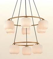 A modern multi-light chandelier with five ribbed glass cylindrical shades and a gold-toned circular metal frame, suspended by black cords against a plain off-white background.
