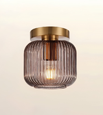 A modern semi-flush mount ceiling light fixture featuring a ribbed, translucent smoky purple glass shade and a brushed brass gold ceiling canopy, shown against a plain off-white background.