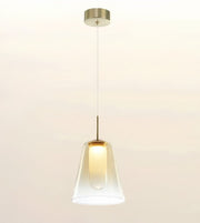 A modern pendant light with a clear glass bell-shaped shade, a warm glowing bulb inside, and a brushed brass metal canopy and rod, suspended against a plain off-white background.