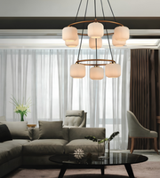 A modern multi-light chandelier with six cylindrical frosted glass shades and a circular bronze metal frame, hanging in a contemporary living room with a gray sectional sofa and large windows.