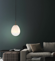 A modern teardrop-shaped pendant light with a glowing frosted glass shade and a brass-toned top cap, hanging in a dark living room above a light gray sofa.