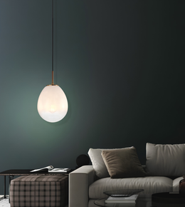 A modern teardrop-shaped pendant light with a glowing frosted glass shade and a brass-toned top cap, hanging in a dark living room above a light gray sofa.