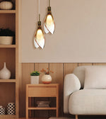 Two modern pendant lights with white leaf-shaped glass shades and bronze metal accents, hanging in a warm, contemporary living room next to a light-colored sofa and wooden side table.