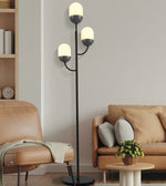 A three-light floor lamp with a matte black metal frame and frosted white glass globes stands in a modern living room next to a tan leather chair and sofa.