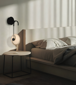A modern wall-mounted black sconce with a circular white shade and curved black arm, installed on a bedroom wall beside a bed and small round side table.