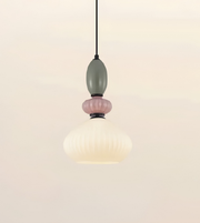 A modern pendant light with a ribbed, bell-shaped frosted white glass shade, accented by a dusty pink glass ring and a muted sage green glass bead, suspended by a thin black cord against a warm off-white background.