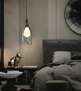 A modern wall-mounted light fixture with a glowing oval bulb and a curved metal frame, installed above a bedside table in a styled bedroom setting.