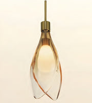 A modern pendant light with a clear glass teardrop-shaped shade featuring organic brown swirls, illuminated with a warm yellow glow, suspended from a brass-colored rod against a light beige background.