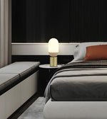 A modern bedroom scene featuring a glowing oval table lamp with a gold cylindrical base on a black nightstand next to a bed with gray bedding and a rust-orange accent pillow.