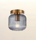 A modern semi-flush mount ceiling light fixture featuring a ribbed, translucent smoky gray glass shade and a brushed gold metal canopy, shown against a soft beige gradient background.