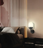 A modern wall-mounted sconce with a glowing white oval shade and a matte dark gray or black base, mounted on a beige wall in a styled bedroom setting next to a bed.