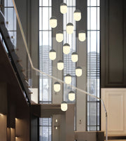 A modern multi-light pendant chandelier with multiple frosted glass dome bulbs suspended at varying heights by curved metal arms, shown hanging in a contemporary indoor staircase lobby.