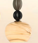 A close-up of a modern pendant light with a rounded, marbled amber glass shade and a sculptural stem featuring stacked matte black and dark gray spherical elements.