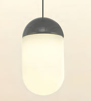 A modern capsule-shaped pendant light with a frosted white cylindrical shade and a glossy dark gray top cap, suspended by a thin black cord against a warm off-white background.