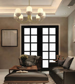 A modern living room interior featuring a multi-light chandelier with white cylindrical glass shades and a gold-toned frame, hanging above a dark leather sofa and wooden coffee table in front of black-framed glass doors.