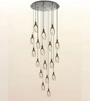 A modern multi-light chandelier with a circular brushed metal ceiling canopy, multiple hanging rods, and twelve leaf-shaped clear glass shades containing warm Edison bulbs, shown against a plain light beige background.