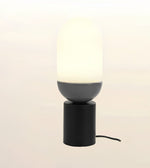 A modern table lamp with a glowing frosted oval shade, a matte dark gray spherical middle section, and a matte black cylindrical base, shown against a soft light beige background.