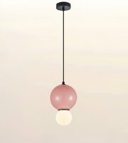 A modern pendant light with a matte black ceiling canopy and cord, featuring a large dusty pink glass globe shade with a smaller white globe bulb hanging below, set against a soft beige gradient background.