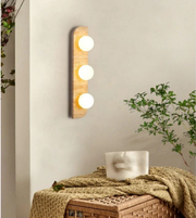 Wooden wall light fixture with three bulbs on a neutral wall with a plant and woven basket in the foreground.