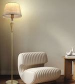 A modern floor lamp with a beige fabric drum shade and a gold-toned metal stand, shown lit in a minimalist room next to a curved white armchair and a small wooden side table.