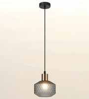 A modern single pendant light with a textured, faceted gray glass shade, a brass-finished metal collar, and a black suspension rod and ceiling canopy, shown against a soft beige gradient background.