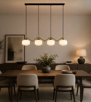 A modern multi-light linear pendant chandelier with four glowing white cylindrical shades and gold-toned geometric accents, hanging over a dining table in a warm, contemporary dining room.