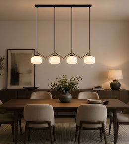 A modern multi-light linear pendant chandelier with four glowing white cylindrical shades and gold-toned geometric accents, hanging over a dining table in a warm, contemporary dining room.